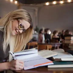 Kobieta w okularach pisze w notatniku, otoczona książkami. Marzy o studiach podyplomowych, na które czeka dofinansowanie.