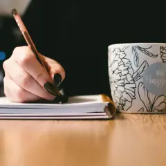 Dłoń pisze długopisem w notatniku, obok kubek z napisem "Be Happy". To inspiracja, jak napisać uzasadnienie wniosku o dofinansowanie.