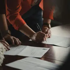 Dłonie podpisują dokumenty na drewnianym stole. Jedna z osób ma na nadgarstku zegarek, a druga nosi bransoletkę. W tle widać fragment pomponu. Klauzula RODO jest ważnym elementem umowy.