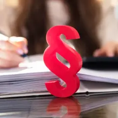 Czerwony paragraf symbolizuje prawne aspekty, gdy firma ogłasza upadłość, co z pracownikami? Tło pokazuje ręce pracujące przy dokumentach i kalkulatorze.