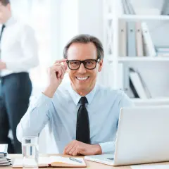 Uśmiechnięty biznesmen poprawia okulary, siedząc przy biurku. Obok laptop, notatnik i szklanka wody. W tle kolega rozmawia przez telefon. Pytanie o zwrot za okulary na kogo faktura?