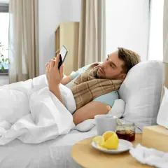Chory mężczyzna w łóżku, owinięty kocem, przegląda telefon. Obok kubek z herbatą, cytryna i miód. To zwolnienie siła wyższa.