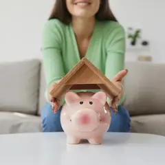 Uśmiechnięta osoba chroni skarbonkę w kształcie świnki drewnianym dachem. Czy na gaz ziemny jest dofinansowanie?