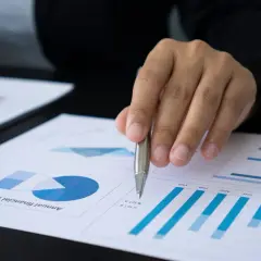 Dłoń z długopisem nad dokumentem z wykresami i danymi finansowymi.