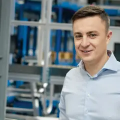 Uśmiechnięty mężczyzna w jasnoniebieskiej koszuli, na tle hali produkcyjnej. To pracownik firmy X KOM, która zajmuje się technologiami.