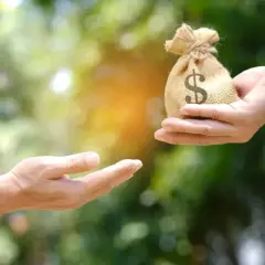 Dwie dłonie, jedna podaje woreczek z pieniędzmi z symbolem dolara, druga wyciągnięta do odbioru.