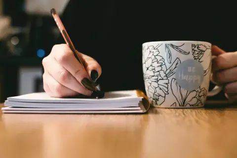 Dłoń pisze długopisem w notatniku, obok kubek z napisem "Be Happy". To inspiracja, jak napisać uzasadnienie wniosku o dofinansowanie.