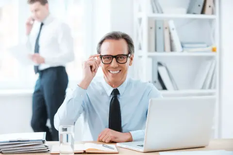Uśmiechnięty biznesmen poprawia okulary, siedząc przy biurku. Obok laptop, notatnik i szklanka wody. W tle kolega rozmawia przez telefon. Pytanie o zwrot za okulary na kogo faktura?