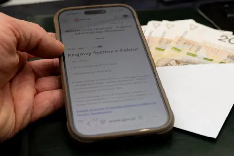 Dłoń trzyma smartfon z informacją o Krajowym Systemie e-Faktur. Obok leżą banknoty, sugerując zwrot VAT.