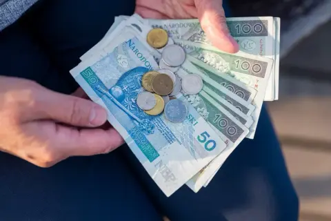 Dłonie trzymają polskie banknoty i monety. Symbolizują one wsparcie finansowe, jak renta rodzinna dla wdowy po ukończeniu 50 lat.