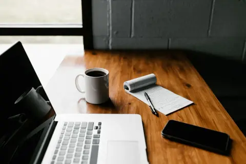 Laptop, kawa, notatnik z długopisem i telefon na drewnianym biurku. Płatnik przygotowuje się do pracy.
