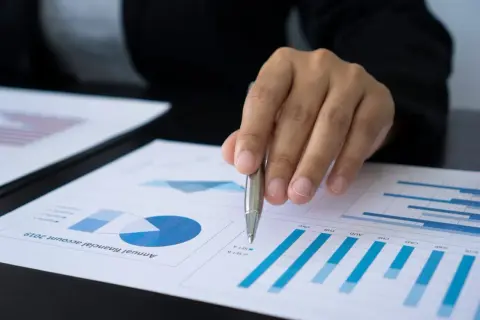 Dłoń z długopisem nad dokumentem z wykresami i danymi finansowymi.