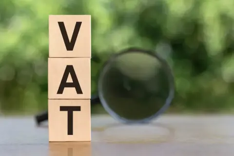 Drewniane klocki z literami VAT ułożone jeden na drugim, obok lupa. Analiza vat naliczony.