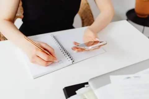 Kobieta notuje w zeszycie, trzymając telefon. Sprawdza, do kiedy można wystawiać faktury za poprzedni miesiąc.