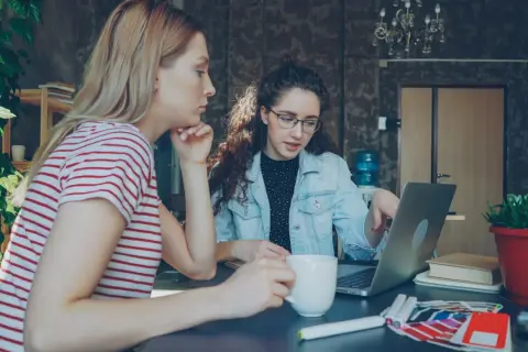 Dwie kobiety pracują nad projektem w biurze. Jedna z nich, z burzą kręconych włosów, wskazuje coś na laptopie. To może być początek ich wspólnej spółki jawnej.