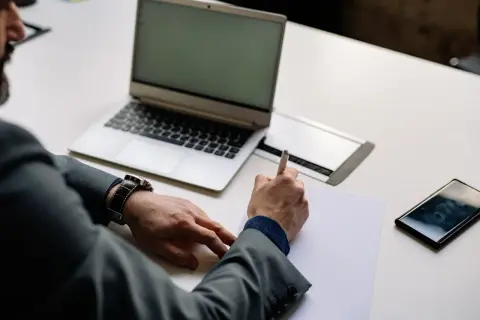 Mężczyzna w garniturze pisze na kartce, obok laptop, telefon i zegarek. Czas na rozliczenie vat-z.