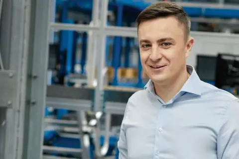 Uśmiechnięty mężczyzna w jasnoniebieskiej koszuli, na tle hali produkcyjnej. To pracownik firmy X KOM, która zajmuje się technologiami.