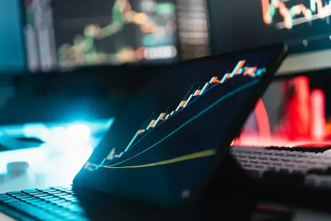 Analiza wykresów giełdowych na ekranie tabletu i monitorów. W tle czerwone i niebieskie światła. To może być inspiracja, jak napisać projekt o dofinansowanie.