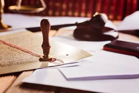 Pieczęć lakowa na dokumencie, młotek sędziowski i waga szalkowa. Symbolizują prawo i porządek, a także to, kto to prokurent – osoba upoważniona do reprezentowania firmy.