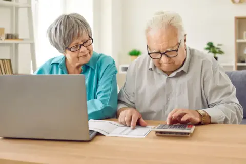 Starsza para przy biurku planuje finanse. Kobieta patrzy na laptopa, mężczyzna liczy na kalkulatorze. To ważny moment przed emerytura wiek.