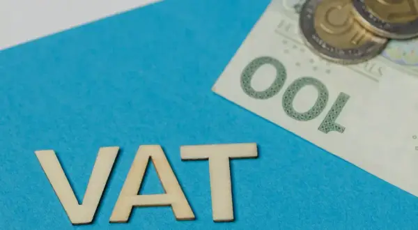 Drewniane litery VAT na niebieskim tle, obok banknot 100 zł i monety. Podatek VAT to ważny element finansów.