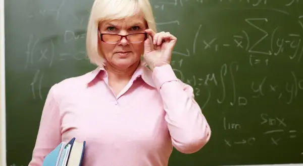 Nauczycielka matematyki z okularami, trzymająca książki, z równaniami na tablicy. Myśli o zasłużonych emeryturach dla nauczycieli.