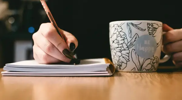 Dłoń pisze długopisem w notatniku, obok kubek z napisem "Be Happy". To inspiracja, jak napisać uzasadnienie wniosku o dofinansowanie.