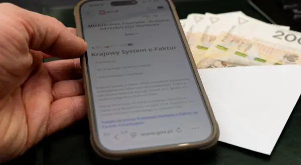 Dłoń trzyma smartfon z informacją o Krajowym Systemie e-Faktur. Obok leżą banknoty, sugerując zwrot VAT.