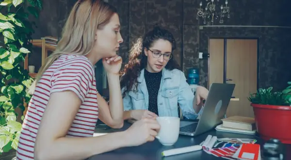 Dwie kobiety pracują nad projektem w biurze. Jedna z nich, z burzą kręconych włosów, wskazuje coś na laptopie. To może być początek ich wspólnej spółki jawnej.