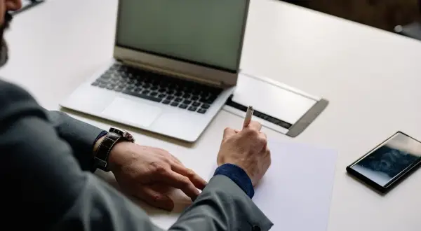 Mężczyzna w garniturze pisze na kartce, obok laptop, telefon i zegarek. Czas na rozliczenie vat-z.