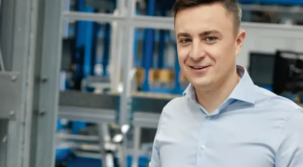 Uśmiechnięty mężczyzna w jasnoniebieskiej koszuli, na tle hali produkcyjnej. To pracownik firmy X KOM, która zajmuje się technologiami.