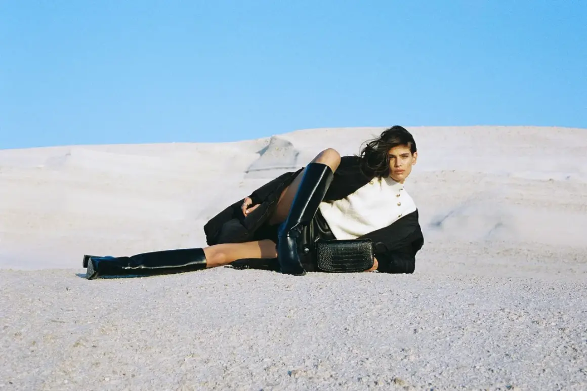 Modelka w kozakach i płaszczu Amisu, z torebką w dłoni, pozuje na tle pustynnego krajobrazu.