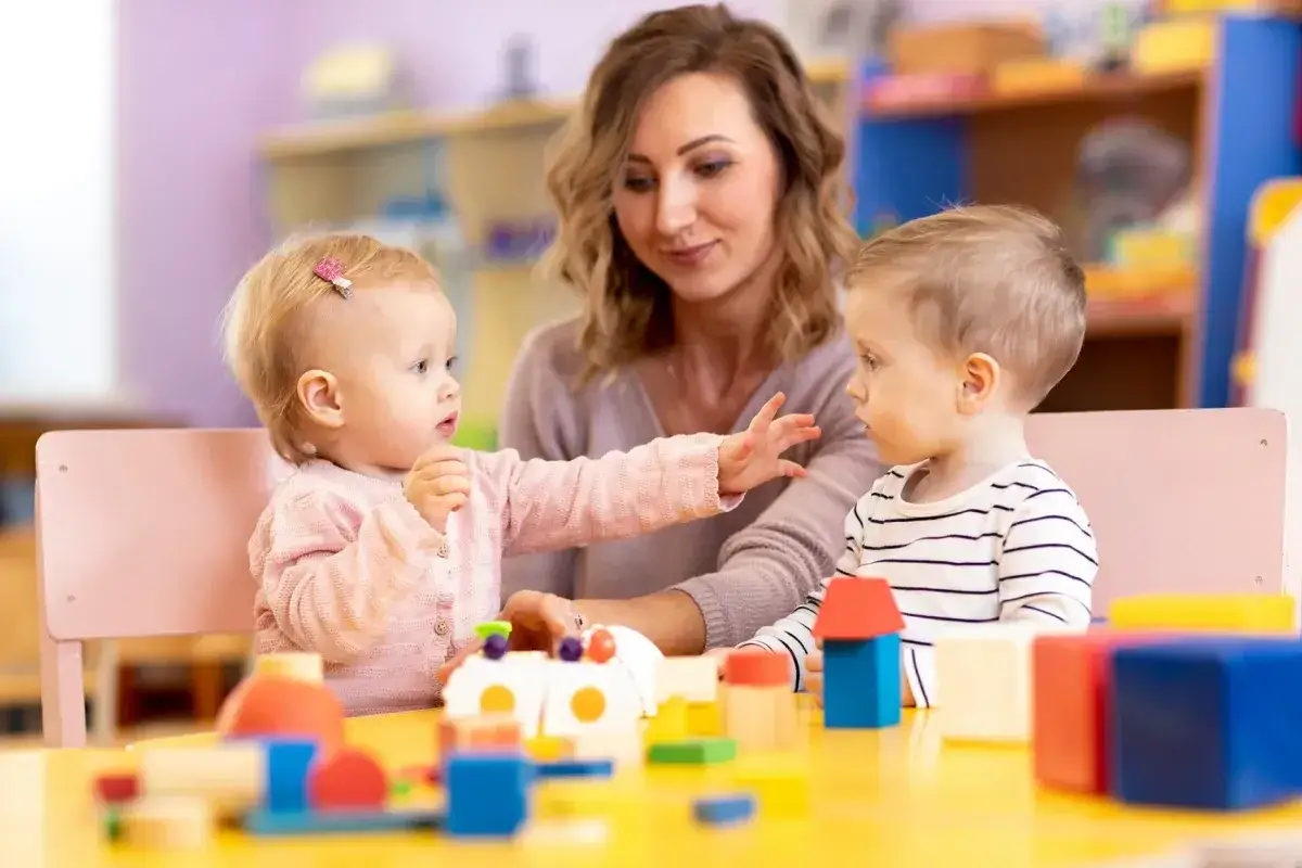Dwie maluchy bawią się klockami, a opiekunka patrzy z uśmiechem. To obrazek z przedszkola, gdzie można dowiedzieć się, jak zdobyć dofinansowanie do przedszkola.