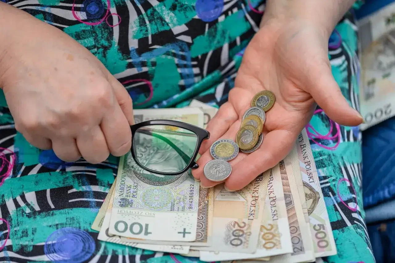Dłonie trzymają okulary i garść monet na tle polskich banknotów. Czy to wystarczy na życie, gdy czeka renta socjalna?