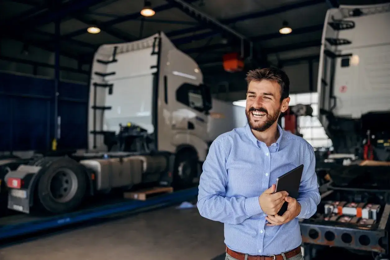 Uśmiechnięty mężczyzna z tabletem w ręku, stojący w warsztacie z ciężarówkami. Planujesz, jak założyć firmę transportową?
