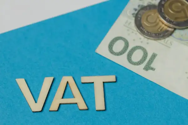 Drewniane litery VAT na niebieskim tle, obok banknot 100 zł i monety. Podatek VAT to ważny element finansów.