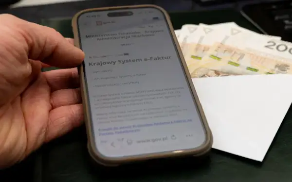 Dłoń trzyma smartfon z informacją o Krajowym Systemie e-Faktur. Obok leżą banknoty, sugerując zwrot VAT.