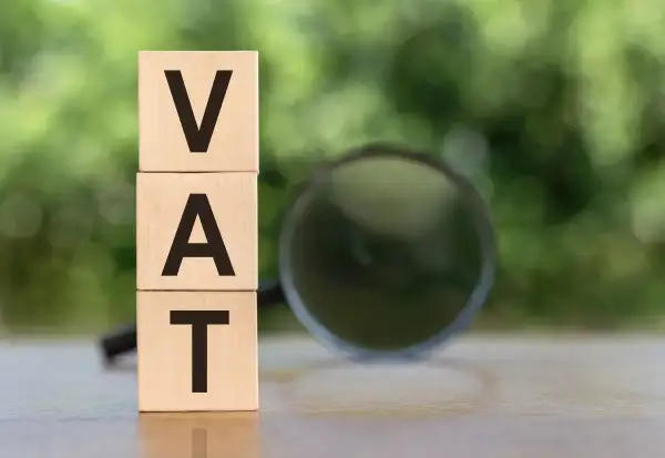 Drewniane klocki z literami VAT ułożone jeden na drugim, obok lupa. Analiza vat naliczony.