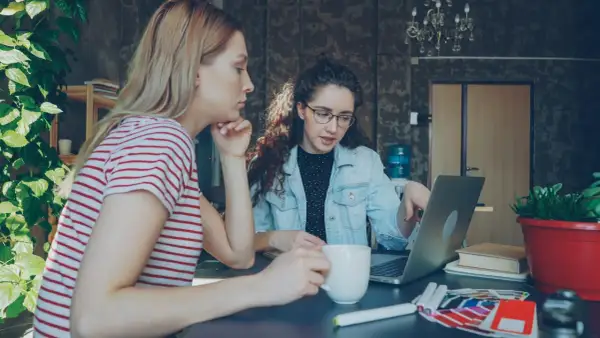 Dwie kobiety pracują nad projektem w biurze. Jedna z nich, z burzą kręconych włosów, wskazuje coś na laptopie. To może być początek ich wspólnej spółki jawnej.